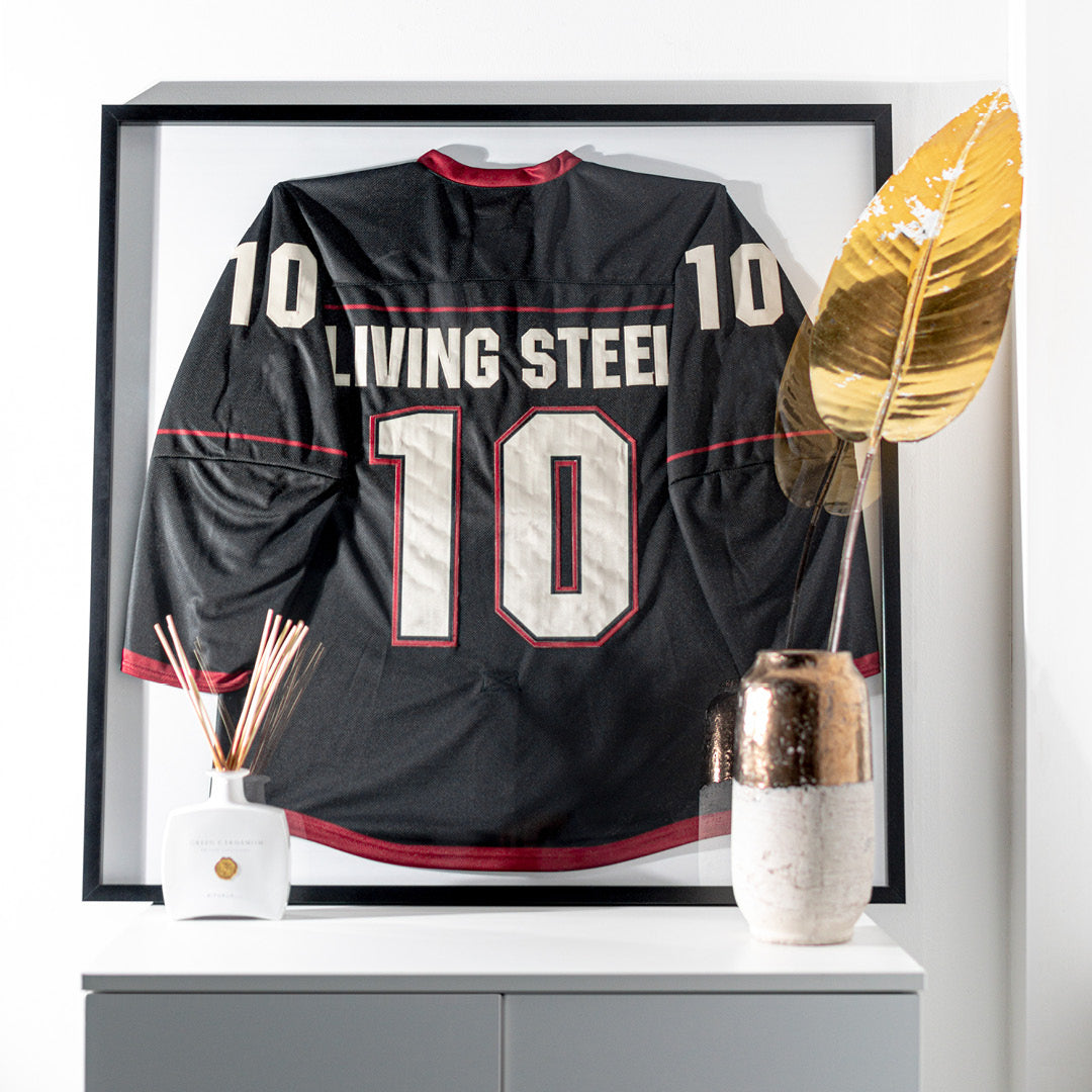 Living.Steel Trikotrahmen mit schwarzem Aluminiumprofil und weißer Rückwand – stilvolle Präsentation eines schwarzen Eishockeytrikots in moderner Wohnumgebung mit dekorativen Elementen.