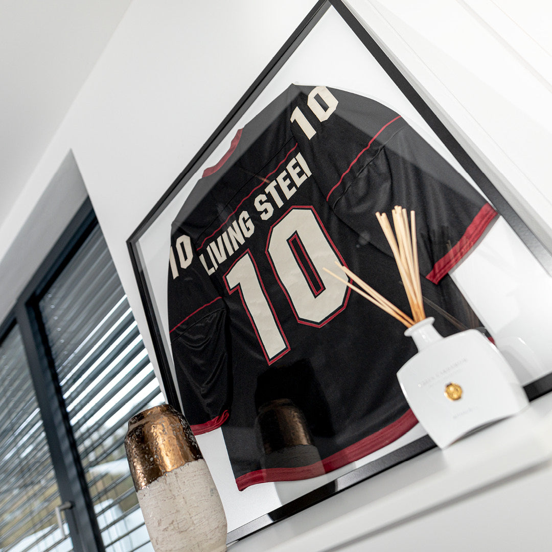Eishockeytrikot im Living.Steel Trikotrahmen – schwarze Aluminiumfassung mit weißer Rückwand, perfekt in Szene gesetzt mit Tageslicht und Deko für maximale Wirkung im Raum.