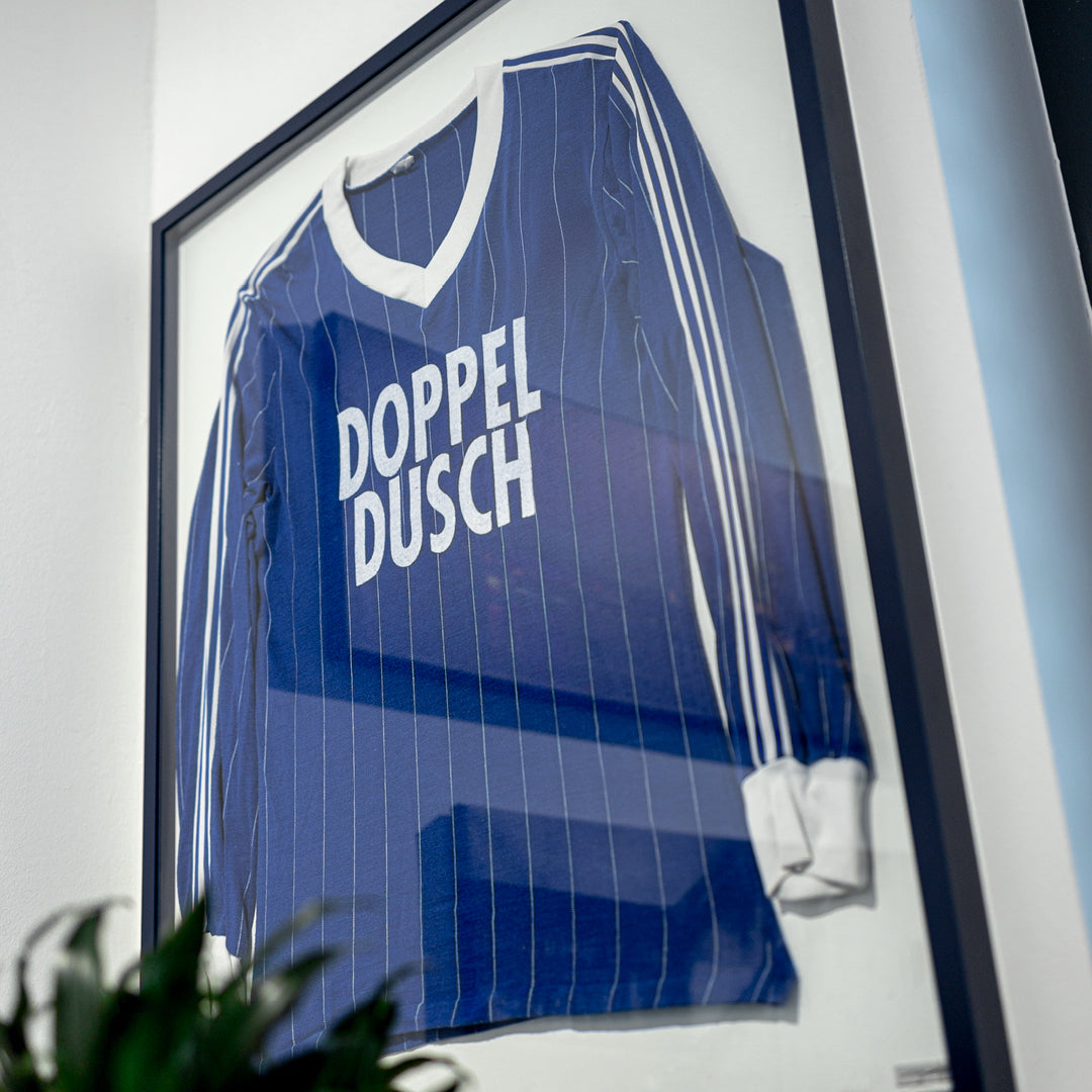Living.Steel Trikotrahmen mit stahlblauem Aluminiumprofil und weißer Rückwand – hier mit einem klassischen Fußballtrikot im Retro-Stil, stilvoll inszeniert hinter Acrylglas.