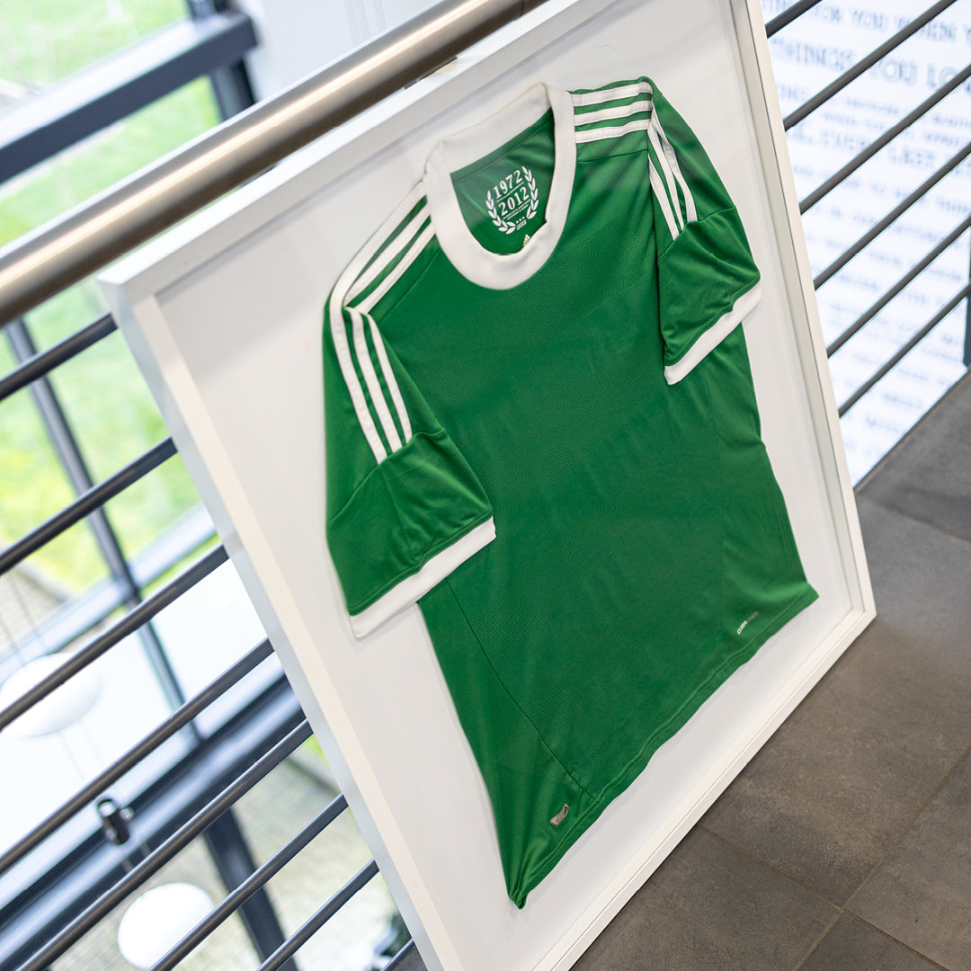 Trikotrahmen von Living.Steel mit weißem Aluminiumrahmen und weißer Rückwand – präsentiert ein grünes Fußballtrikot in moderner Umgebung, ideal für Sammler und Fans.